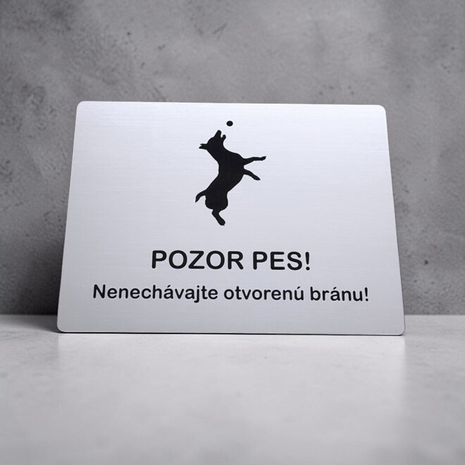 Biela tabuľka so skákajúcim Jack Russell teriérom a nápisom Nenechávajte otvorenú bránu