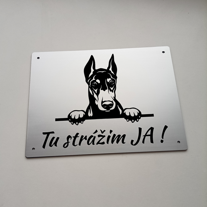 Strieborná tabuľka s vykúkajúcim dobermanom a nápisom Tu strážim ja